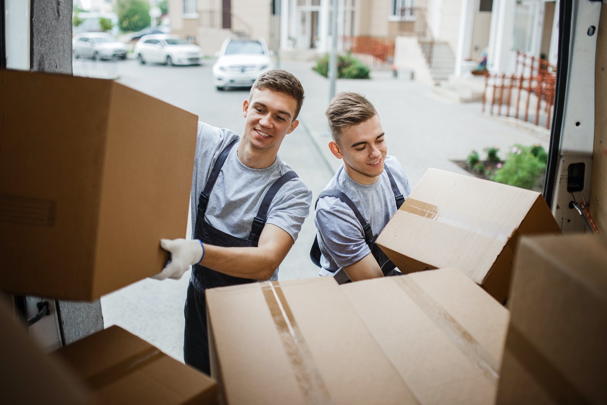 Two young handsome smiling movers wearing uniforms are unloading the van full of boxes. House move